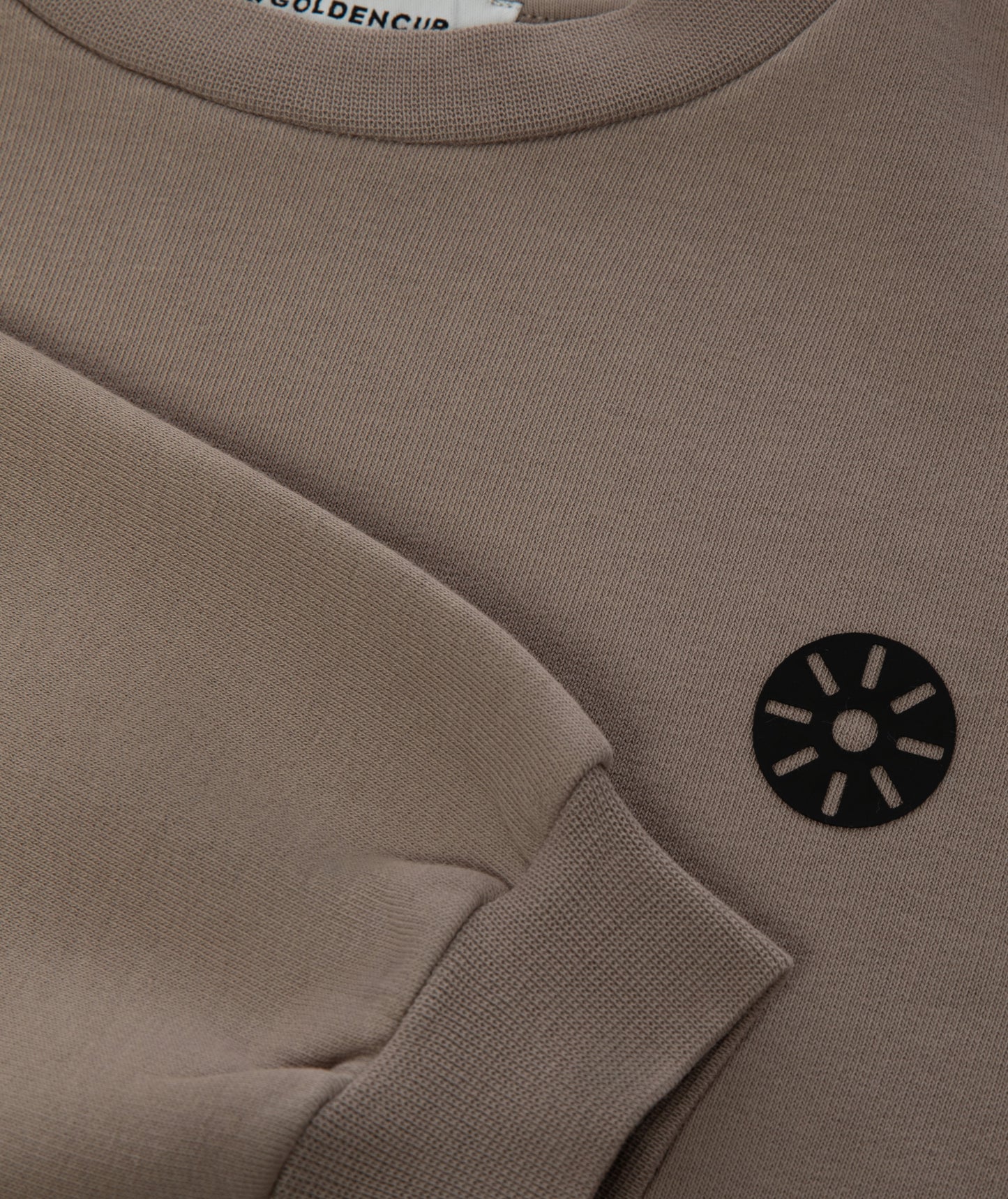 Close-up of Goldencubs black circular logo printed on soft taupe cotton sweatshirt fabric.