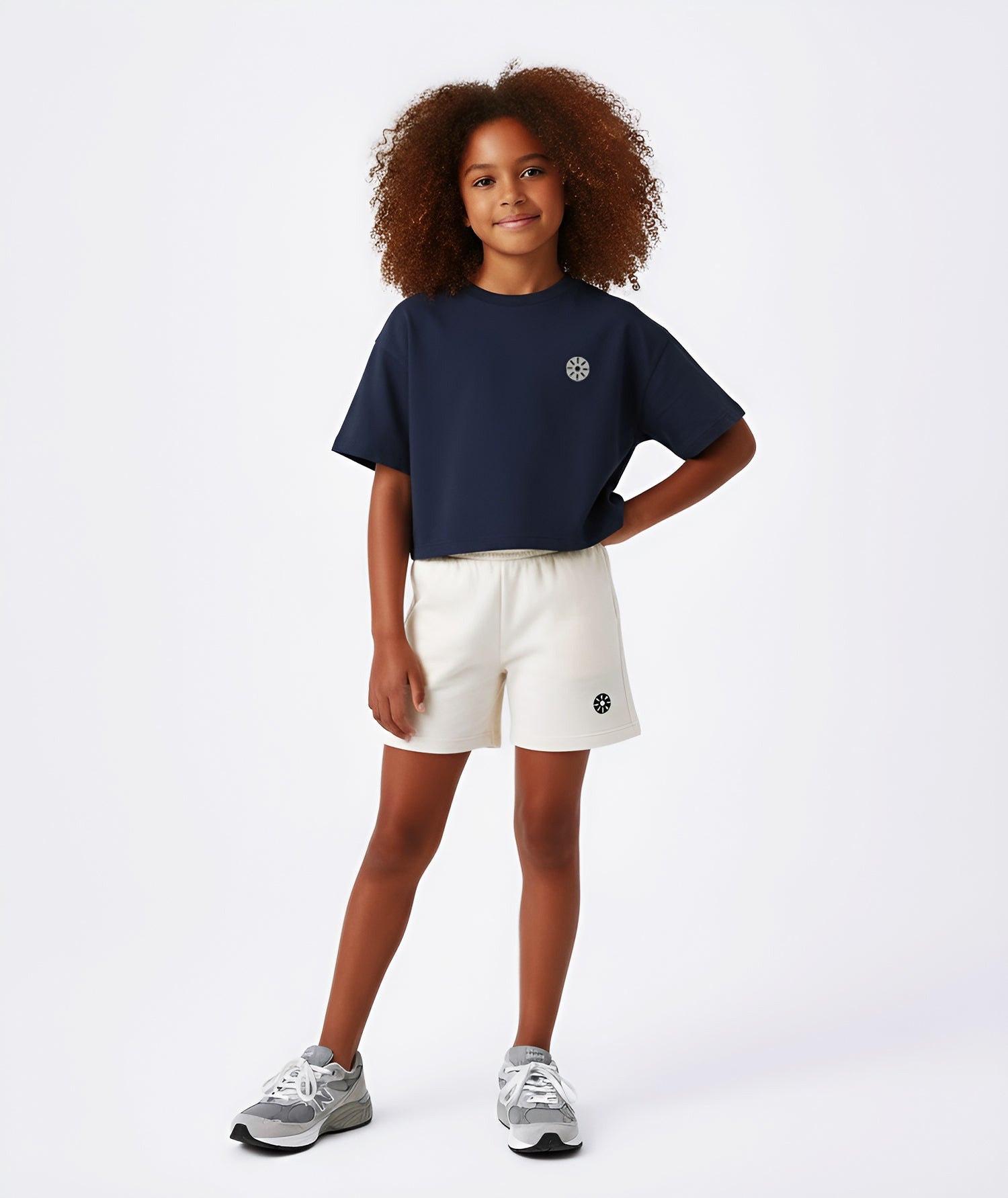 Girl wearing Goldencubs navy cotton cropped T-shirt with gray circular logo, paired with cream shorts and gray sneakers.
