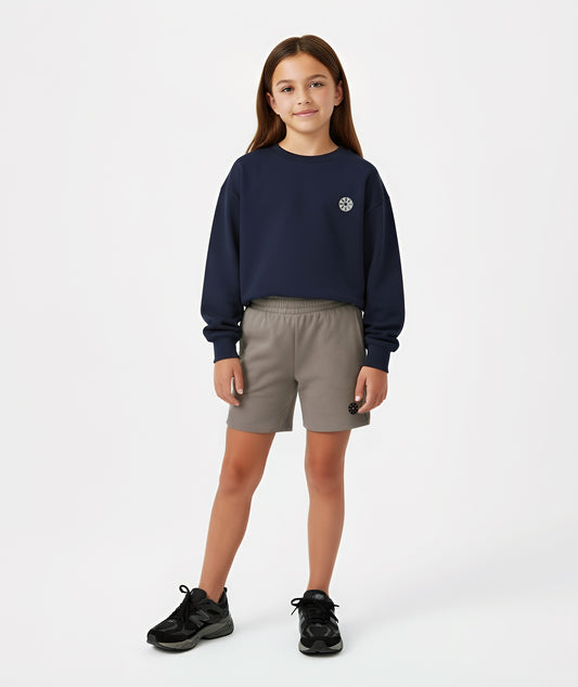 Girl wearing Goldencubs navy cotton sweatshirt with gray circular logo, styled with taupe shorts and black sneakers.