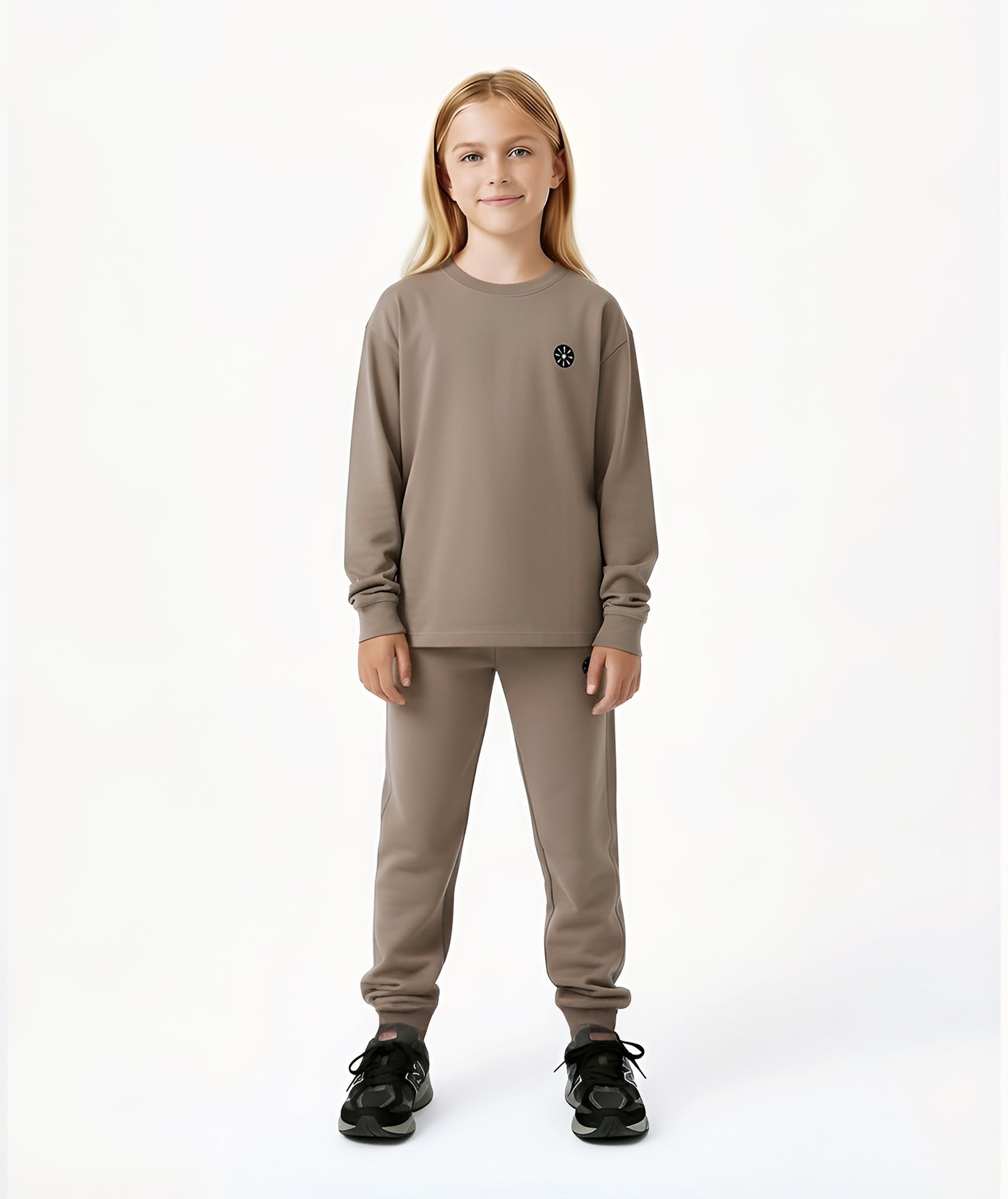Girl wearing Goldencubs taupe cotton long sleeve T-shirt with black circular logo, paired with matching joggers and black sneakers.