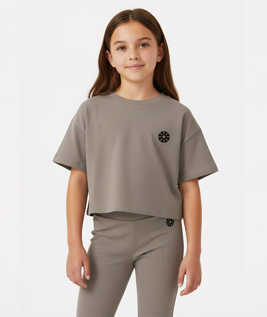 Girl wearing Goldencubs taupe cotton T-shirt with short sleeves and black circular logo, posing against a white background.