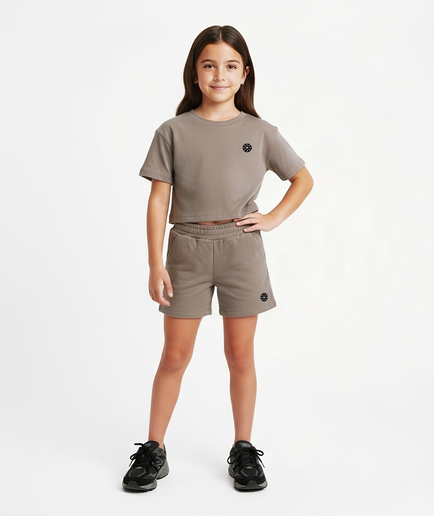Girl wearing Goldencubs taupe cotton cropped T-shirt with black circular logo, styled with matching shorts and black sneakers.